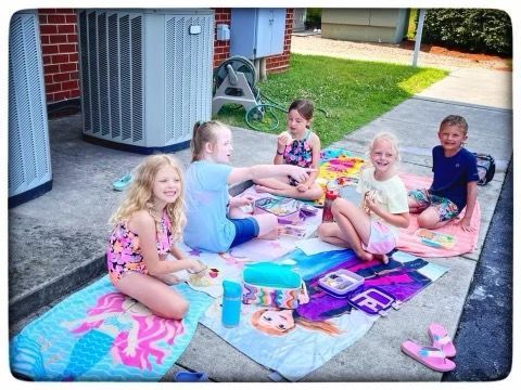 A group of children are sitting on a blanket on the sidewalk.