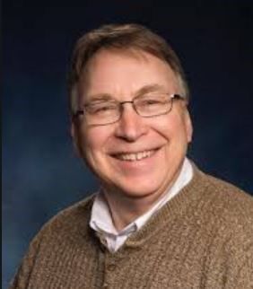 A man wearing glasses and a brown sweater is smiling for the camera.