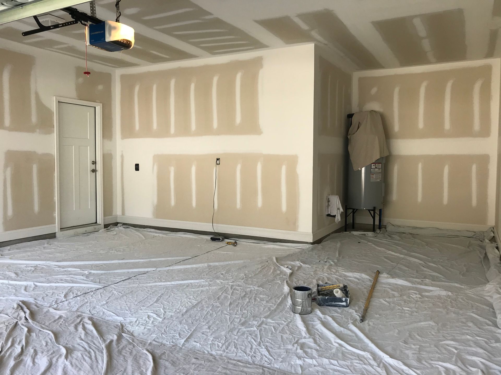 Garage interior with drywall installation in progress. Walls and ceiling are mostly white. Floor is covered with drop cloths.