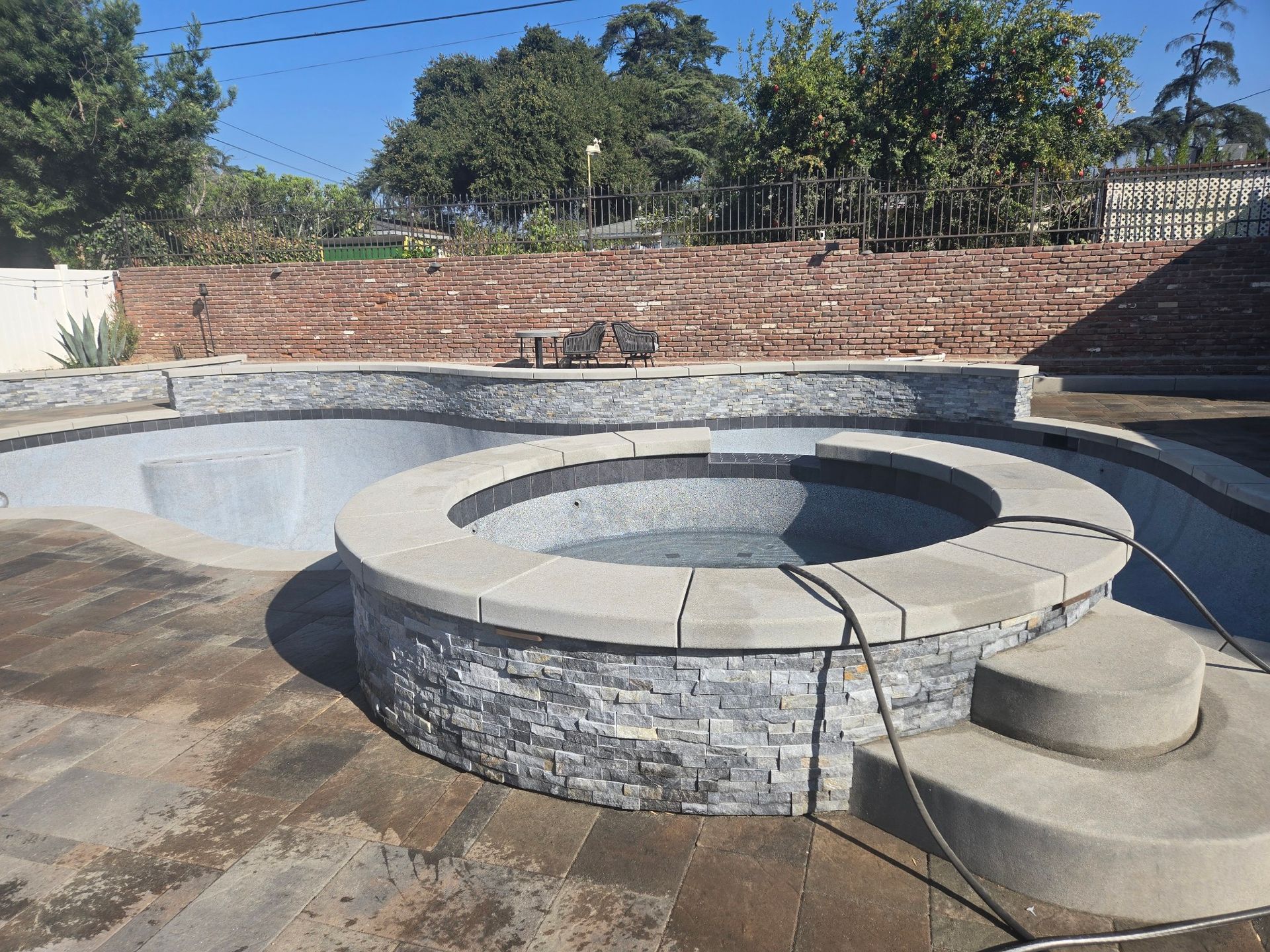 A newly constructed pool and spa with gray stone features and a dark tiled interior, set in a sunny backyard.