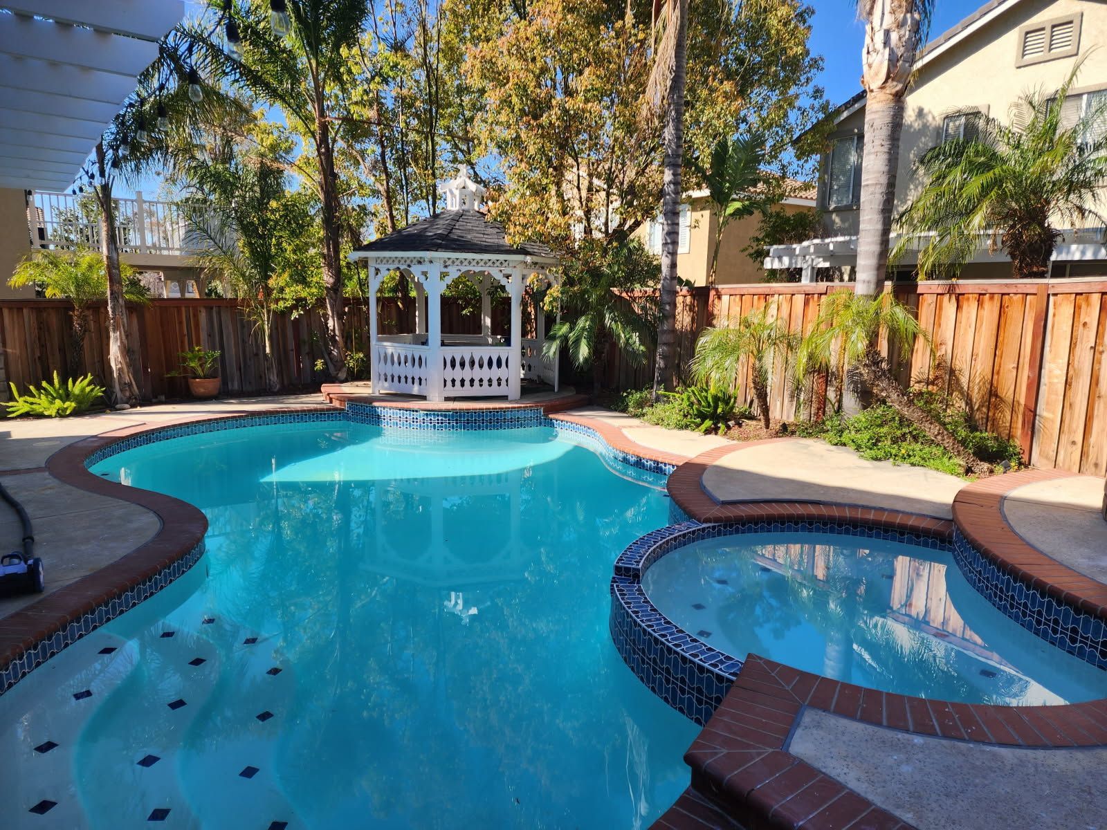 A backyard pool with turquoise water and a built-in hot tub, featuring a gazebo in the background, surrounded by a wooden fence and greenery.