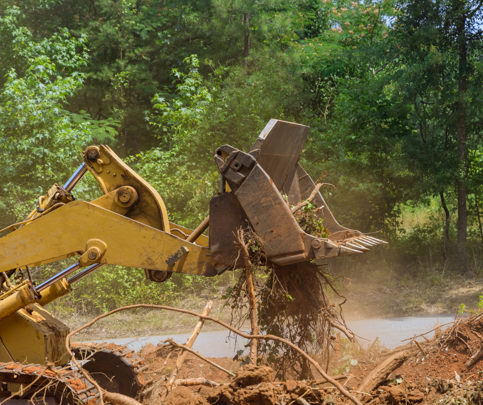 Yellow excavator bucket pulling roots and mud in a wooded, muddy construction site
