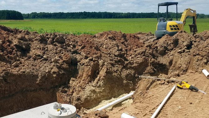 Excavator digging a trench beside white pipes at a construction site, with dirt piles and green fields nearby