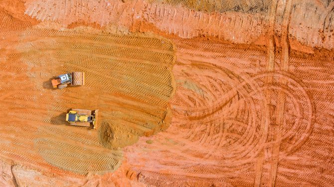 Two construction vehicles on a reddish dirt road near circular tire tracks