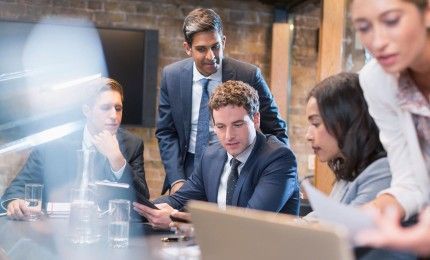 A group of business people are sitting around a table looking at a laptop.