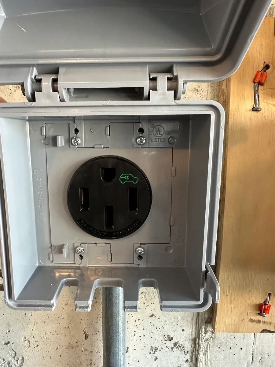 Open gray outdoor electrical outlet box with a black receptacle and exposed wiring on a plywood wall.