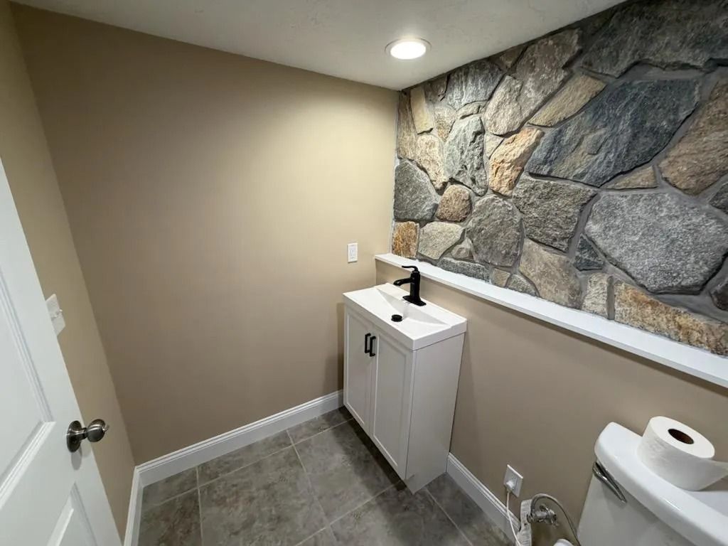 Small bathroom with white vanity, stone accent wall, and toilet paper on a side shelf.