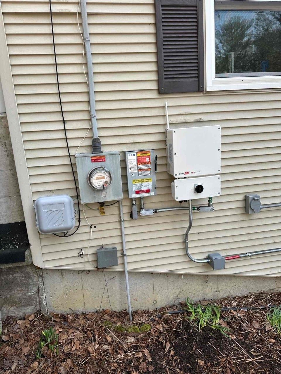 Outdoor house utility meters and electrical boxes mounted on beige siding beside a window.
