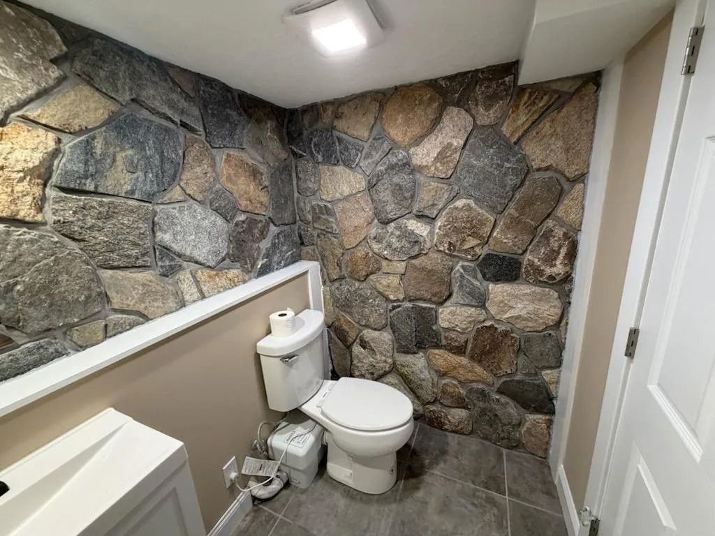 Small restroom with stone accent wall, toilet, and white handrail under a ceiling light.