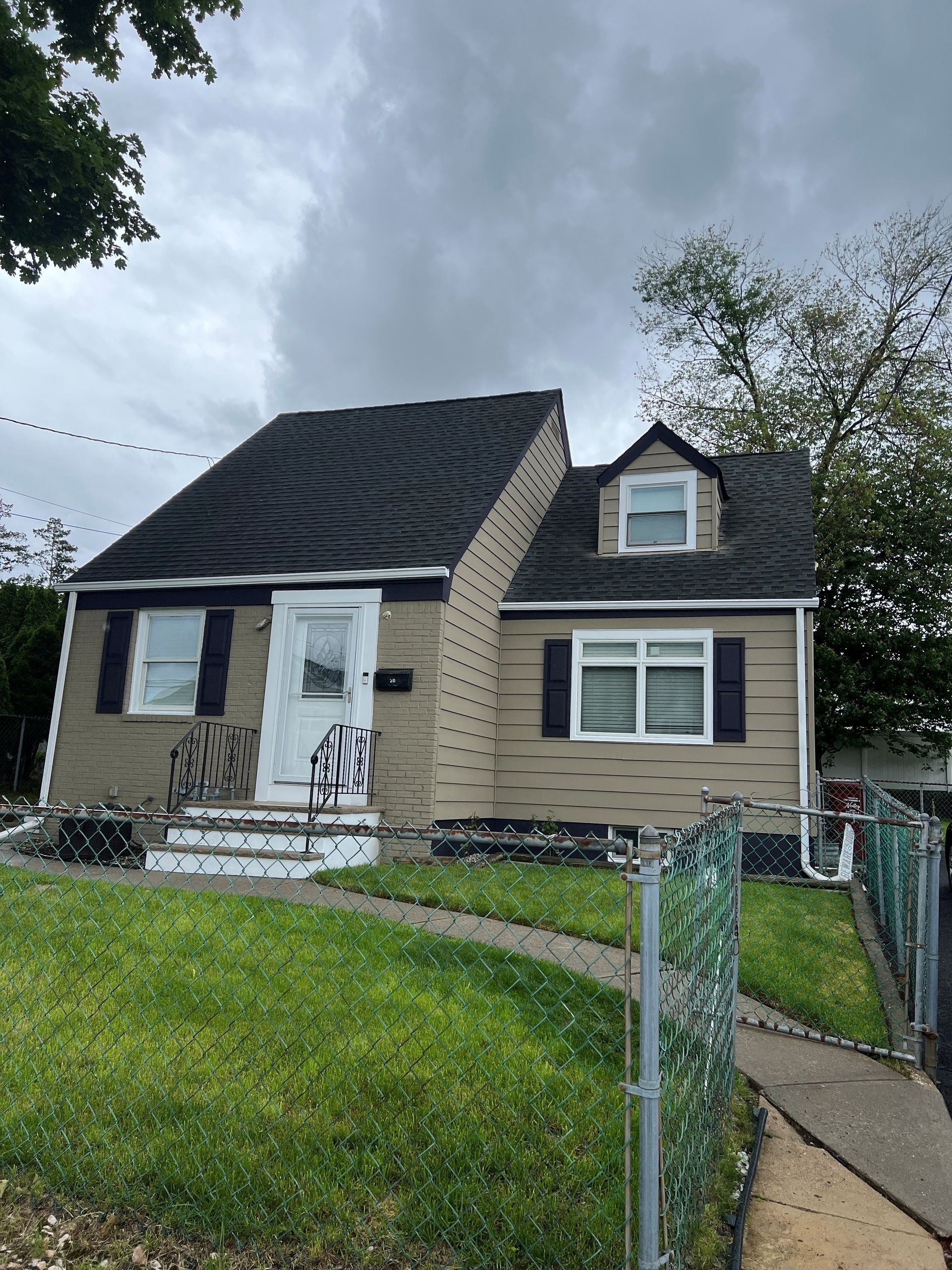 Tan house with black shutters, chimney, and gray roof, green lawn, and chain-link fence.