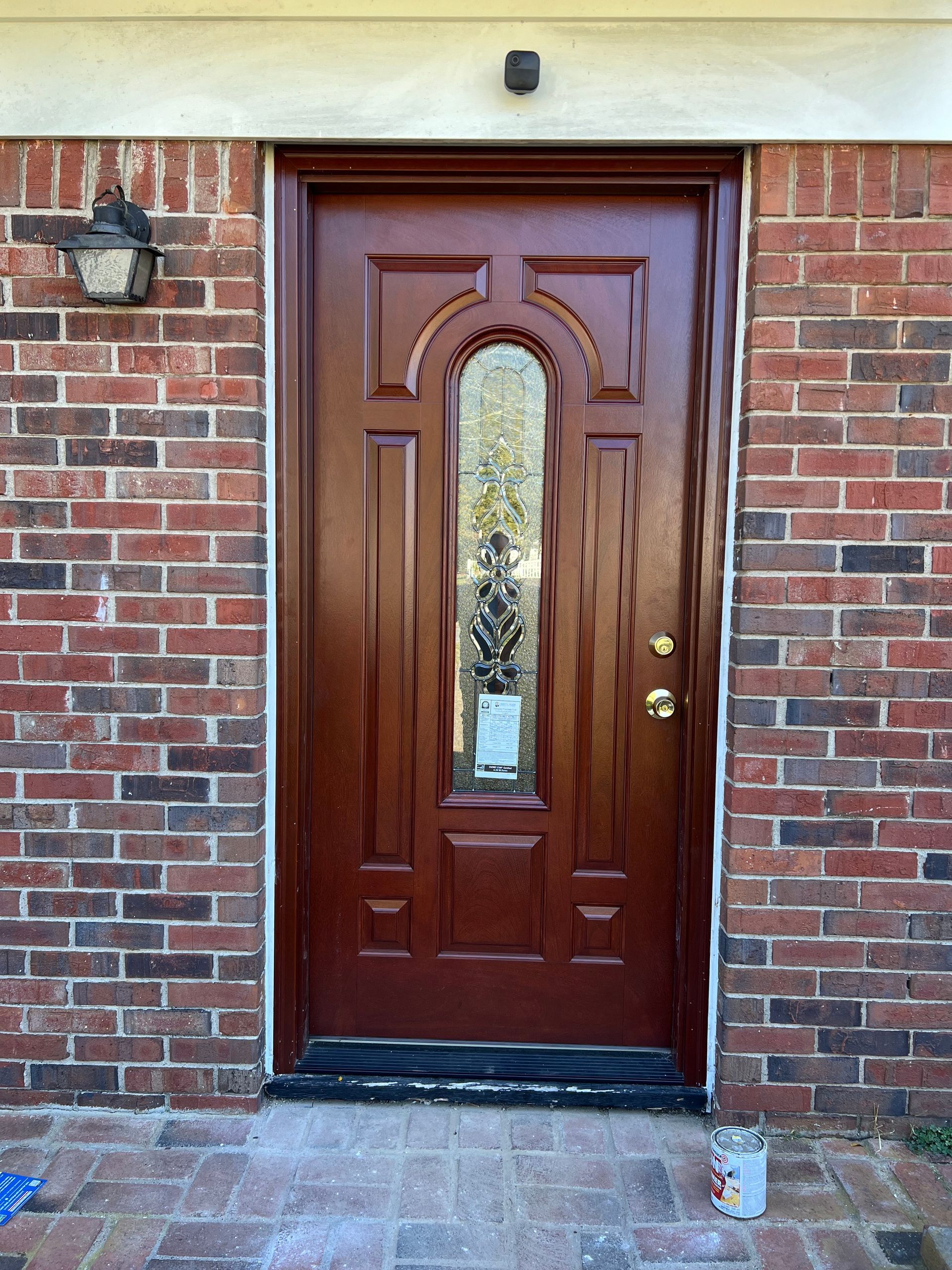 Brown front door with glass panel, surrounded by red brick, and a small black security camera above.