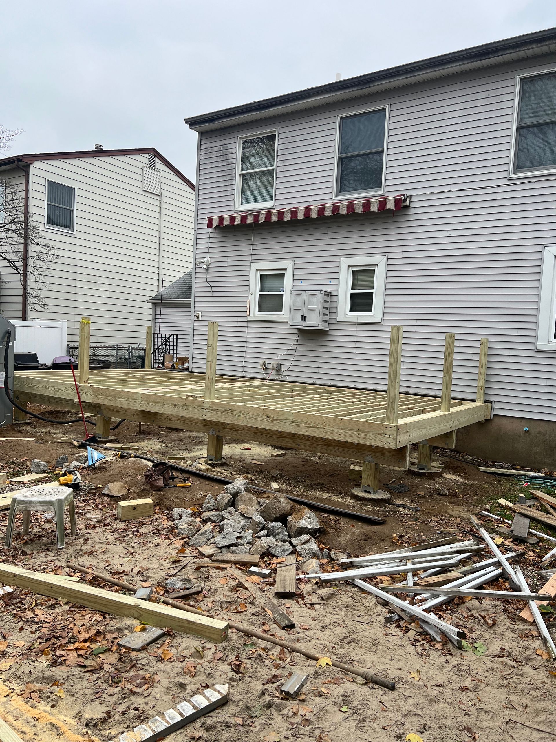 Deck construction in backyard; wooden frame elevated, unfinished with surrounding debris and two-story house.