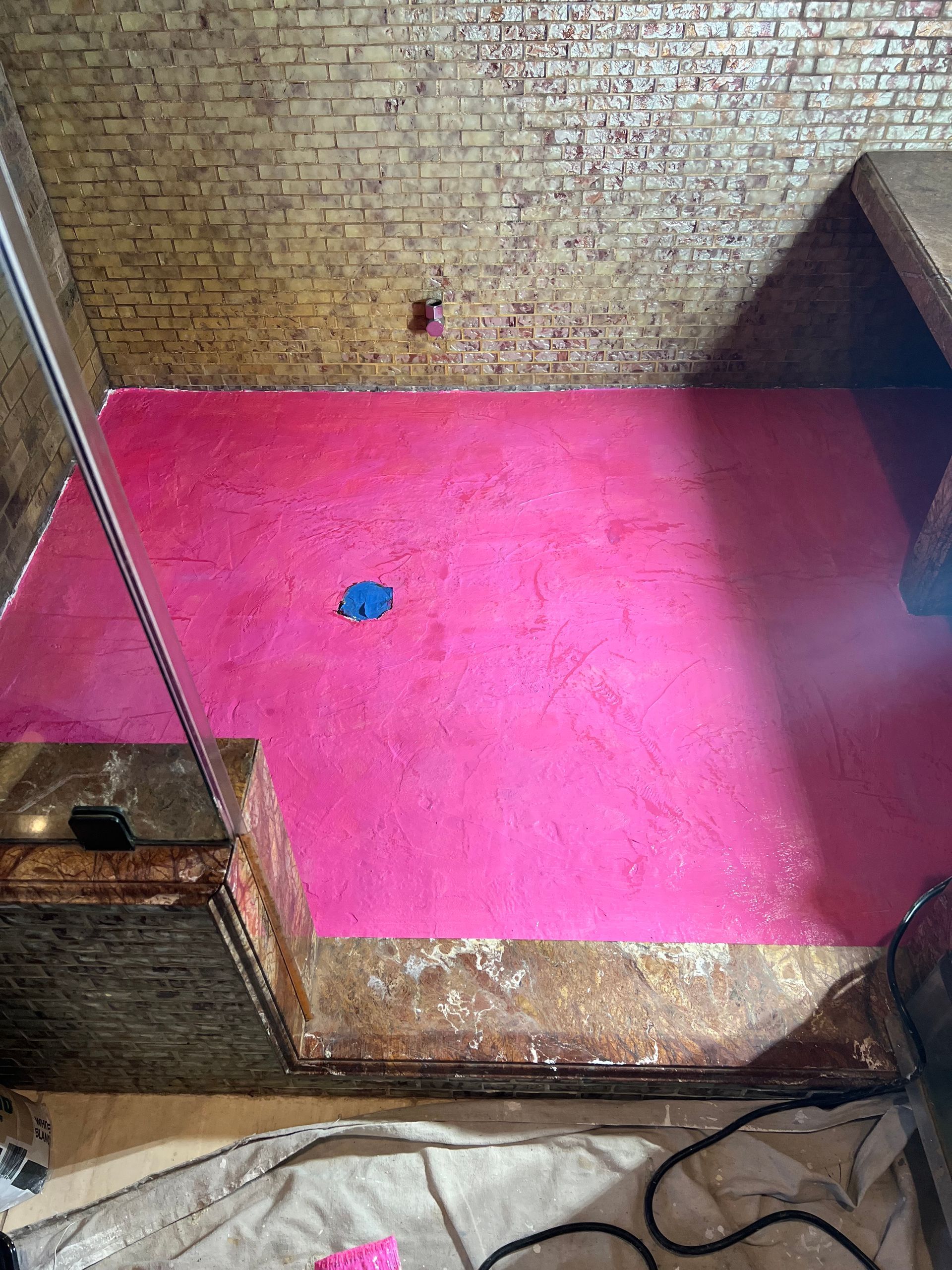 Bright pink painted floor in a brick-walled room. Wooden ledges and a blue drain are visible.