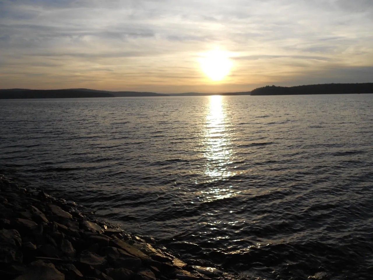 Sunset over a calm lake with golden light reflecting on the water and rocky shoreline in the foreground