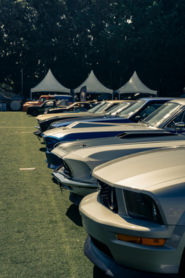 Row of parked classic cars at an outdoor show, with white tents and trees in the background.