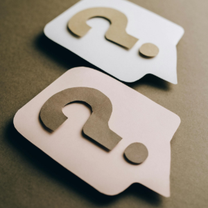 Two paper speech bubbles with question marks, one white and one beige, on a brown background