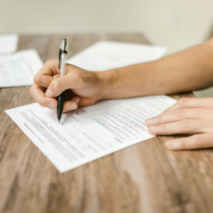 Hands signing a document on a wooden table with papers in the background