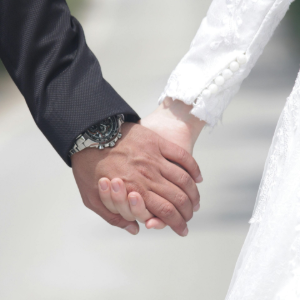 Two people holding hands, one in a dark suit and one in a white dress, with a watch visible.