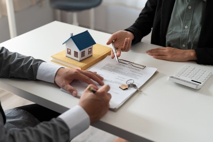 Hands discussing a house model and contract on a white desk with a calculator