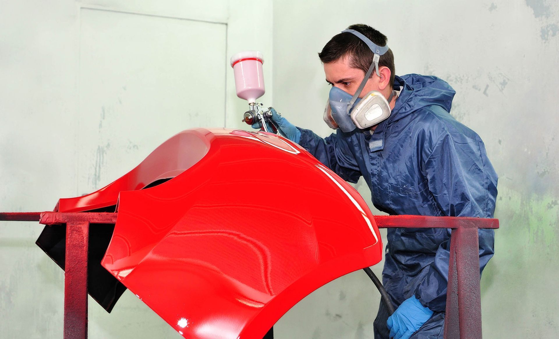 Painter spraying red car hood in a workshop while wearing protective gear and a mask
