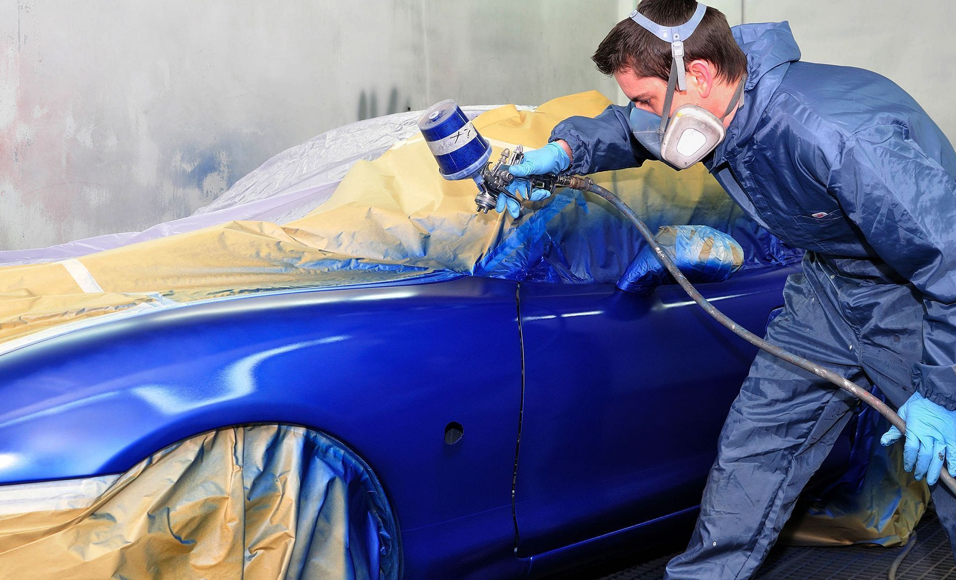Person spray-painting a blue car in a workshop, wearing protective gear and a respirator