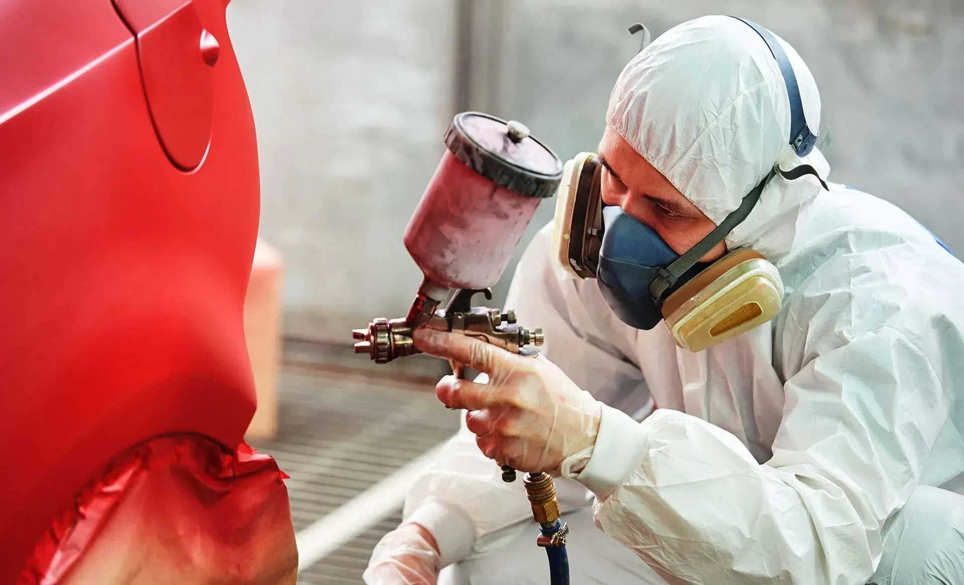 Worker in white protective suit spray-painting a red car door with a spray gun