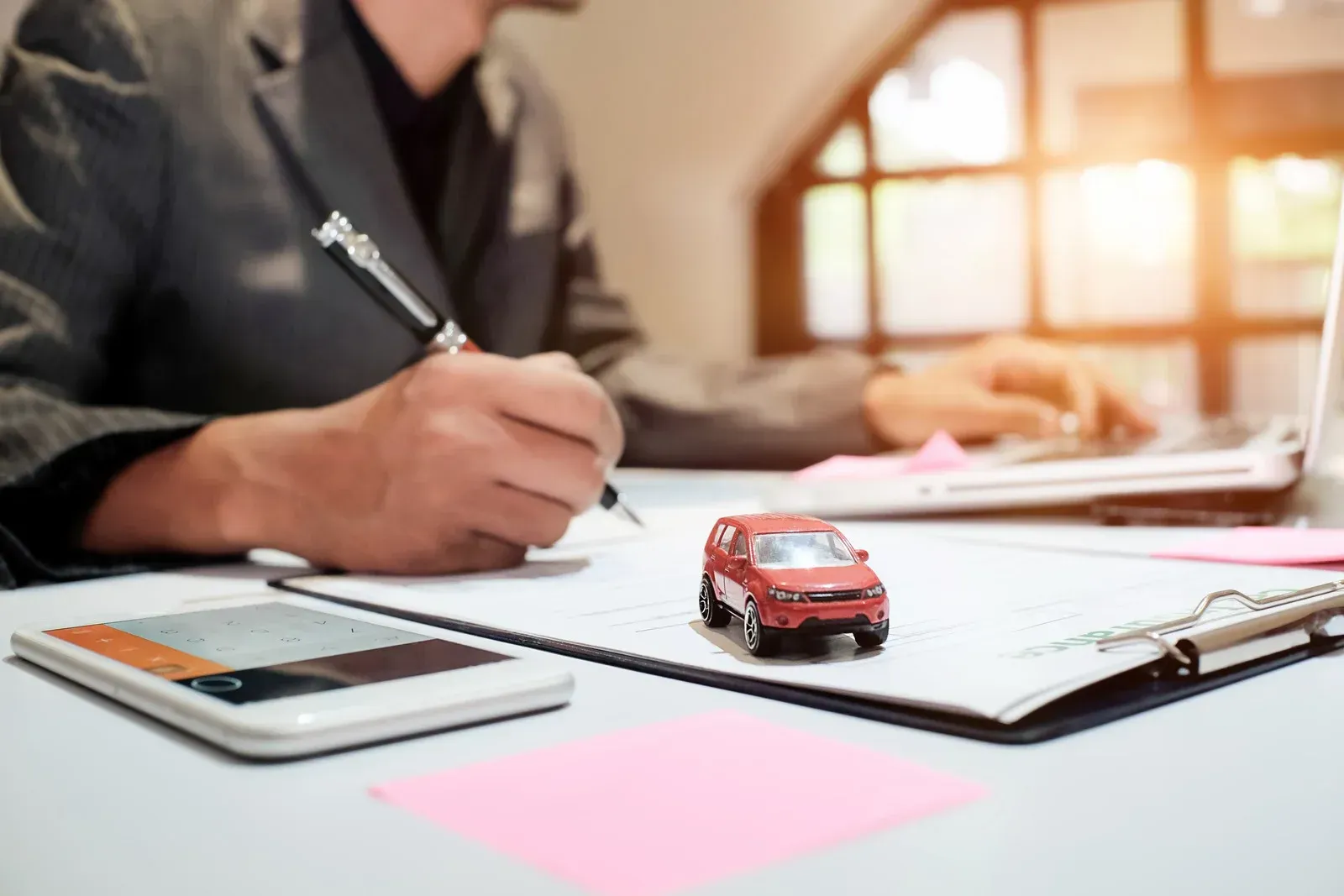 Person in suit writing on document, toy car, calculator, and laptop on a desk.