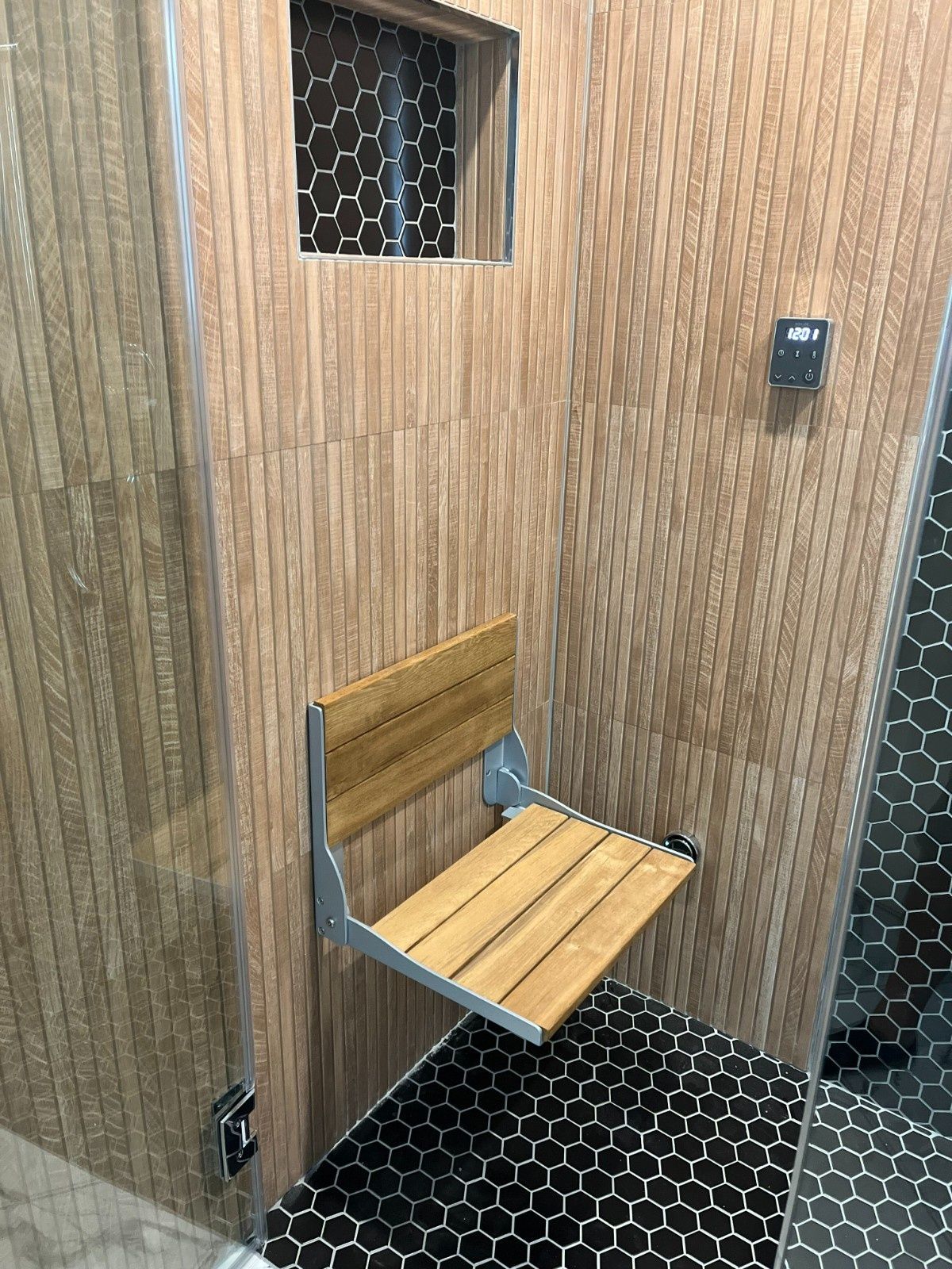 Small wooden restroom stall with a wall-mounted folding bench over black hex tile flooring.