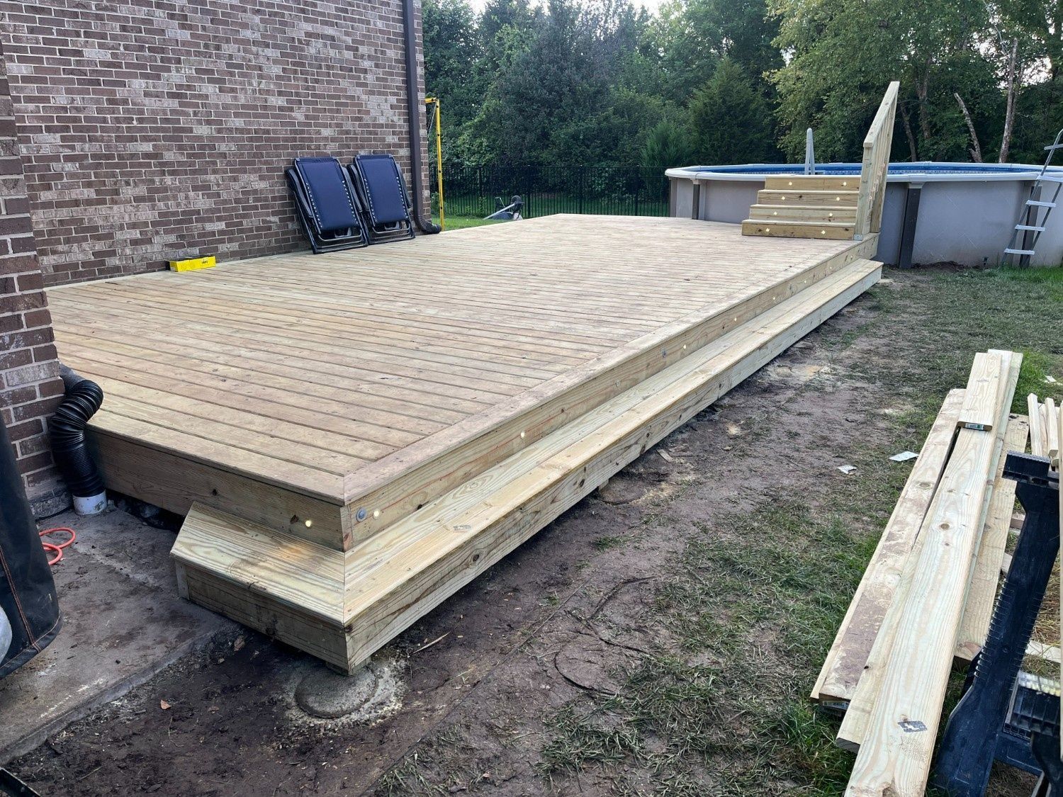 Raised wooden deck platform under construction beside a brick house, with stacked lumber nearby.