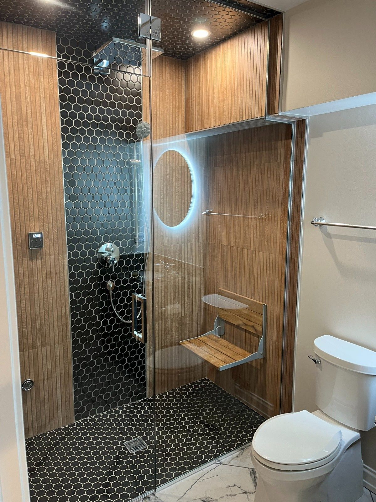 Modern bathroom with black tile shower, wood paneling, round mirror, bench, and toilet