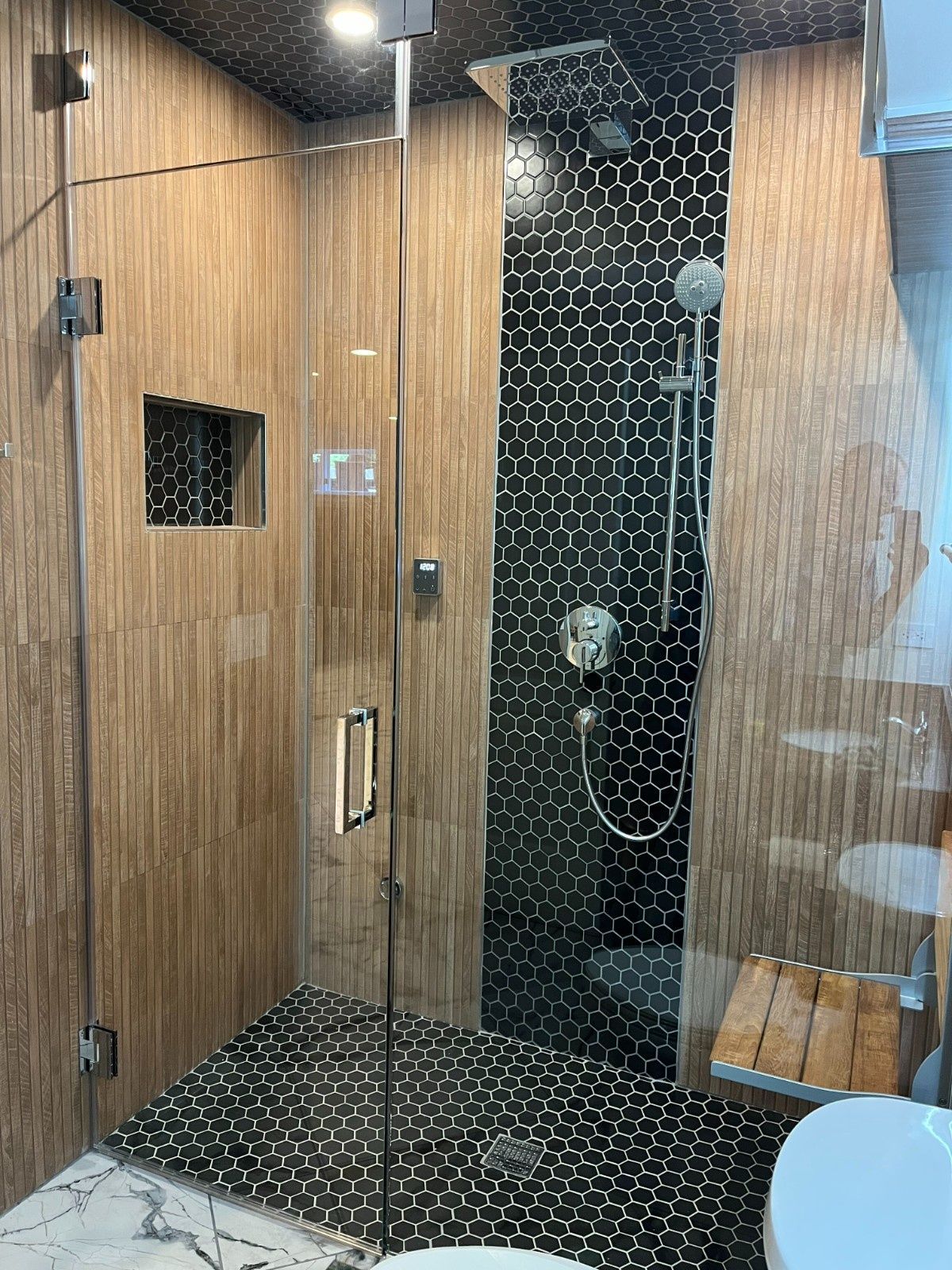 Small modern shower with wood-look walls, black hex tile, glass door, and a showerhead inside.