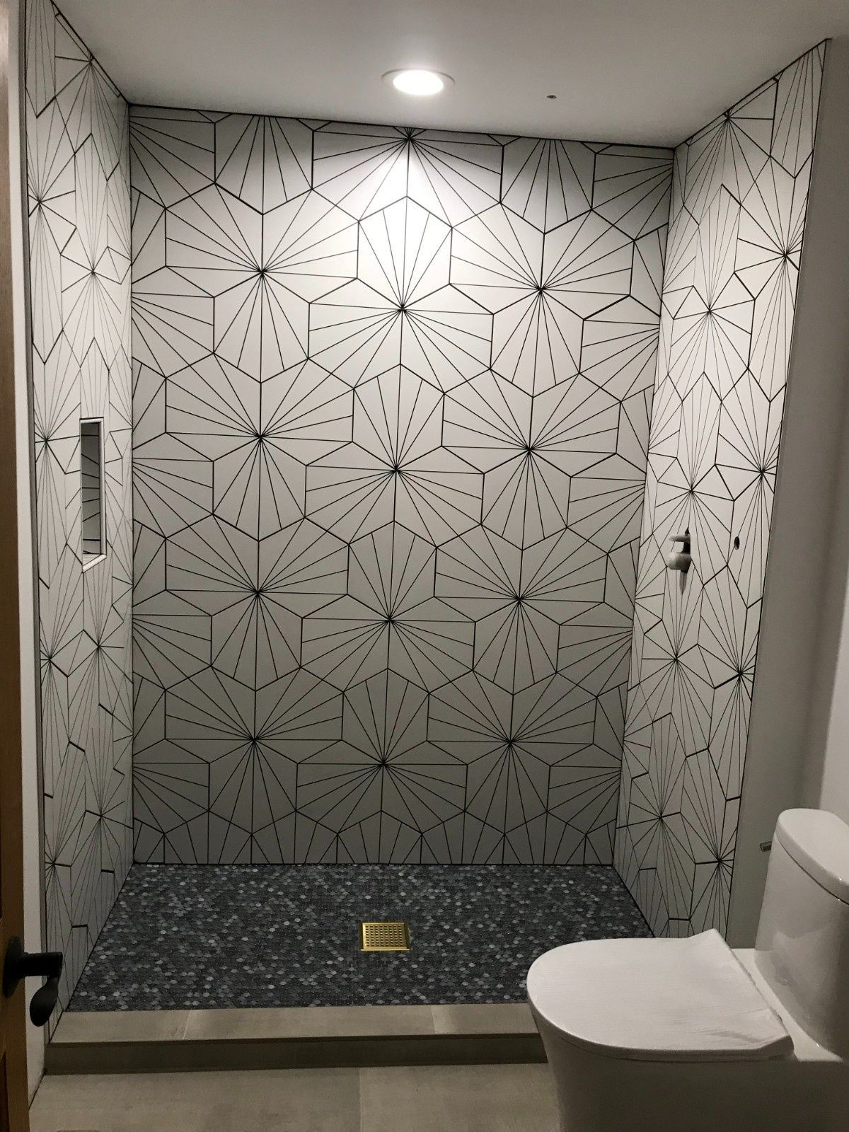 Small tiled bathroom with geometric walls, gray floor, and a white toilet in the foreground.