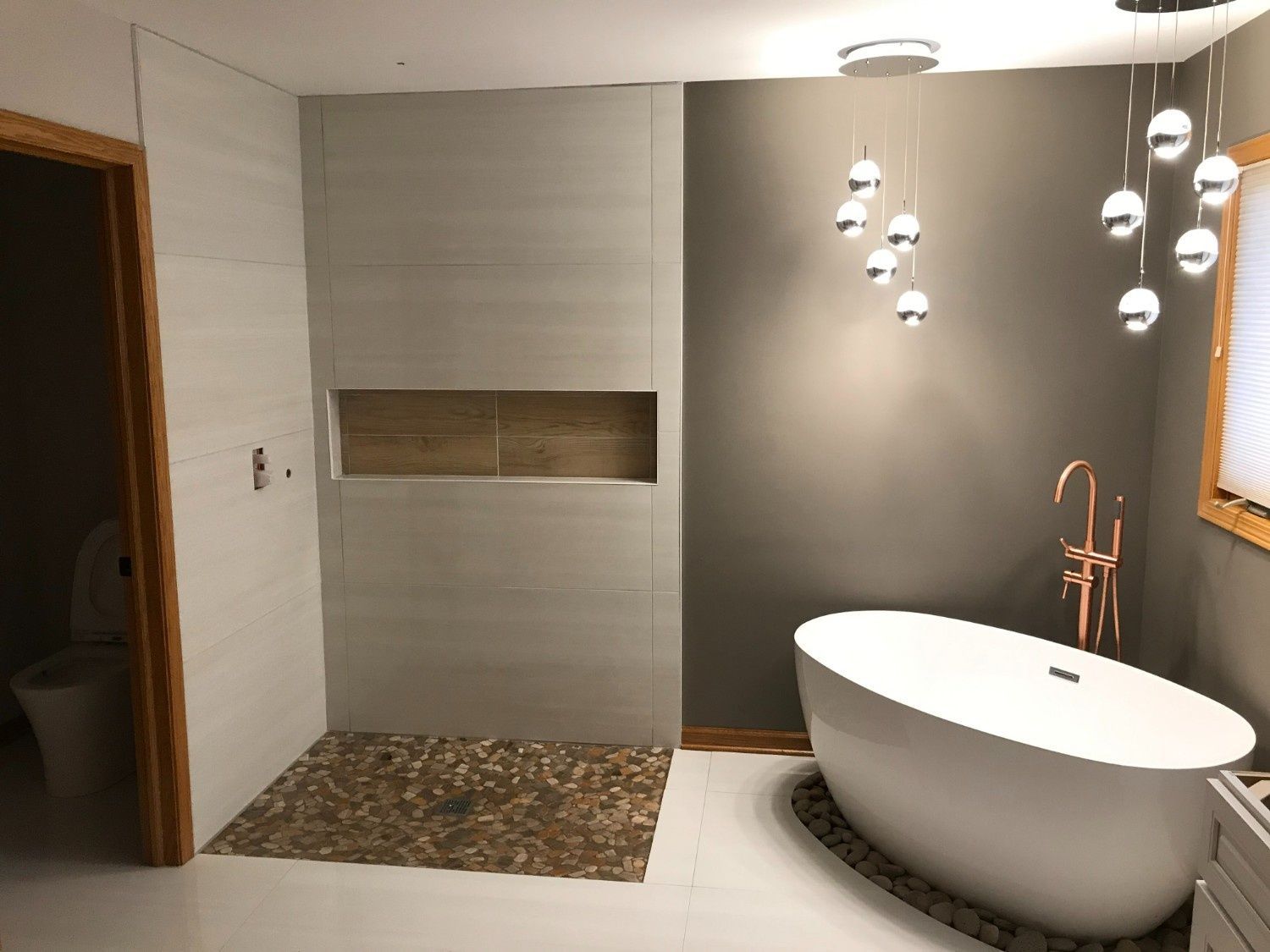 Modern bathroom with oval bathtub, hanging pendant lights, gray wall, and tiled shower niche