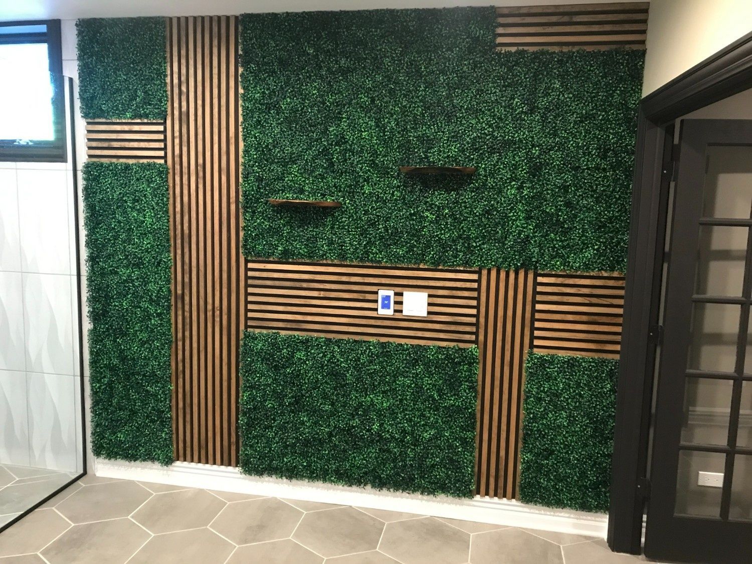Green leafy wall with vertical wood slats, a small central control panel, and a doorway on the right.