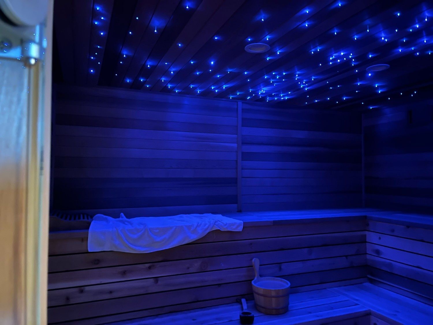 Blue-lit wooden sauna with benches, towel on the wall, bucket on the floor, and starry ceiling lights