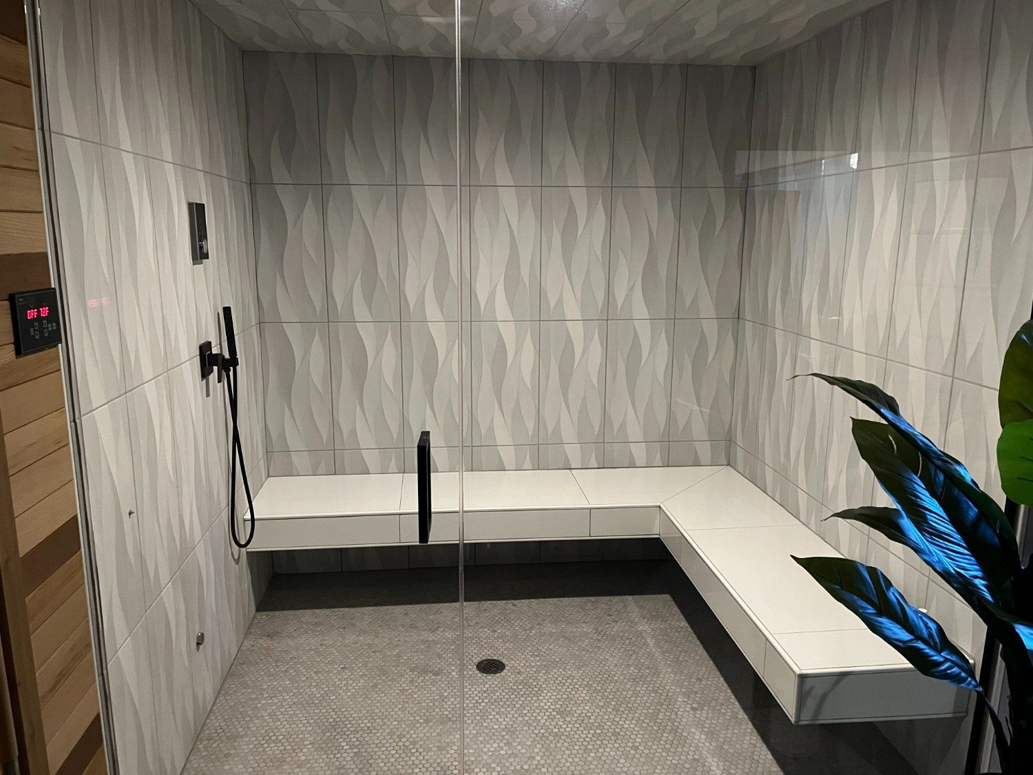 Empty modern shower stall with gray tile walls, built-in bench, handheld shower, and floor drain