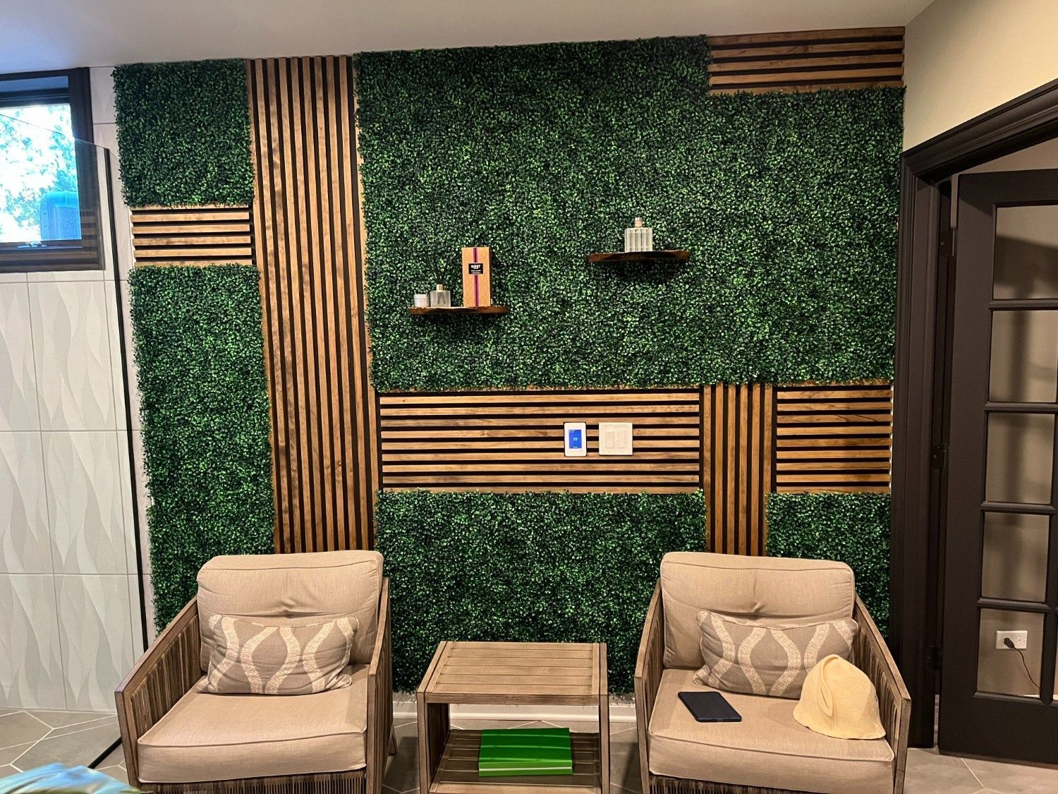 Modern lounge with green leafy wall panels, wooden slats, and two beige armchairs beside a small table.
