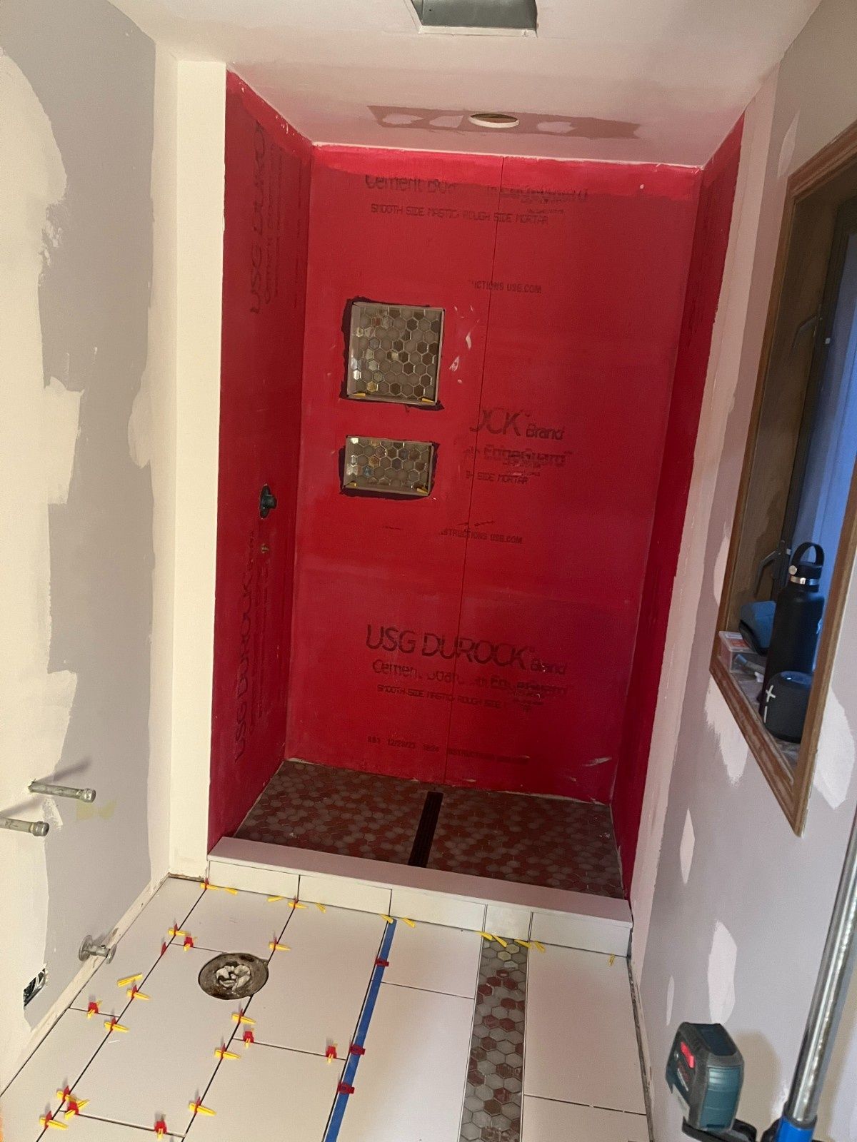 Red-walled shower stall under renovation with tools and tiled floor in a narrow bathroom corridor