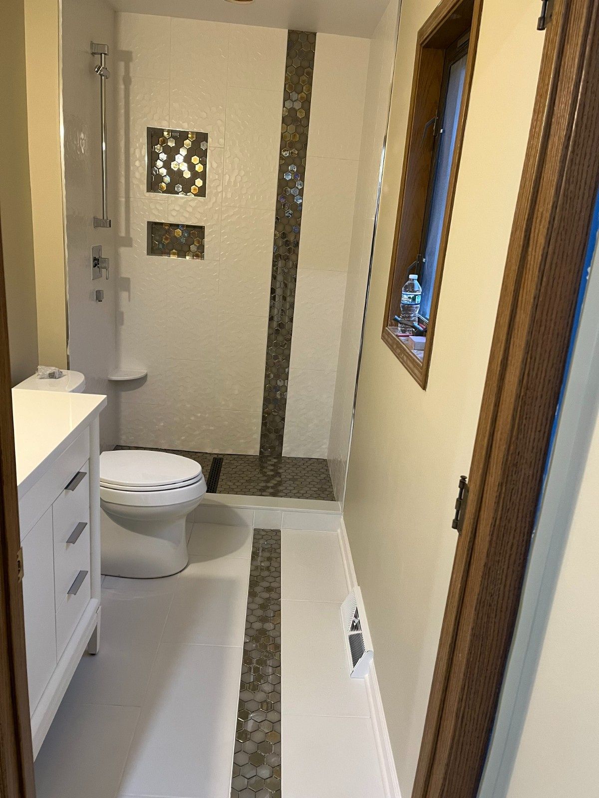 Narrow modern bathroom with toilet, vanity, tub, mosaic tile, and window beside the shower area