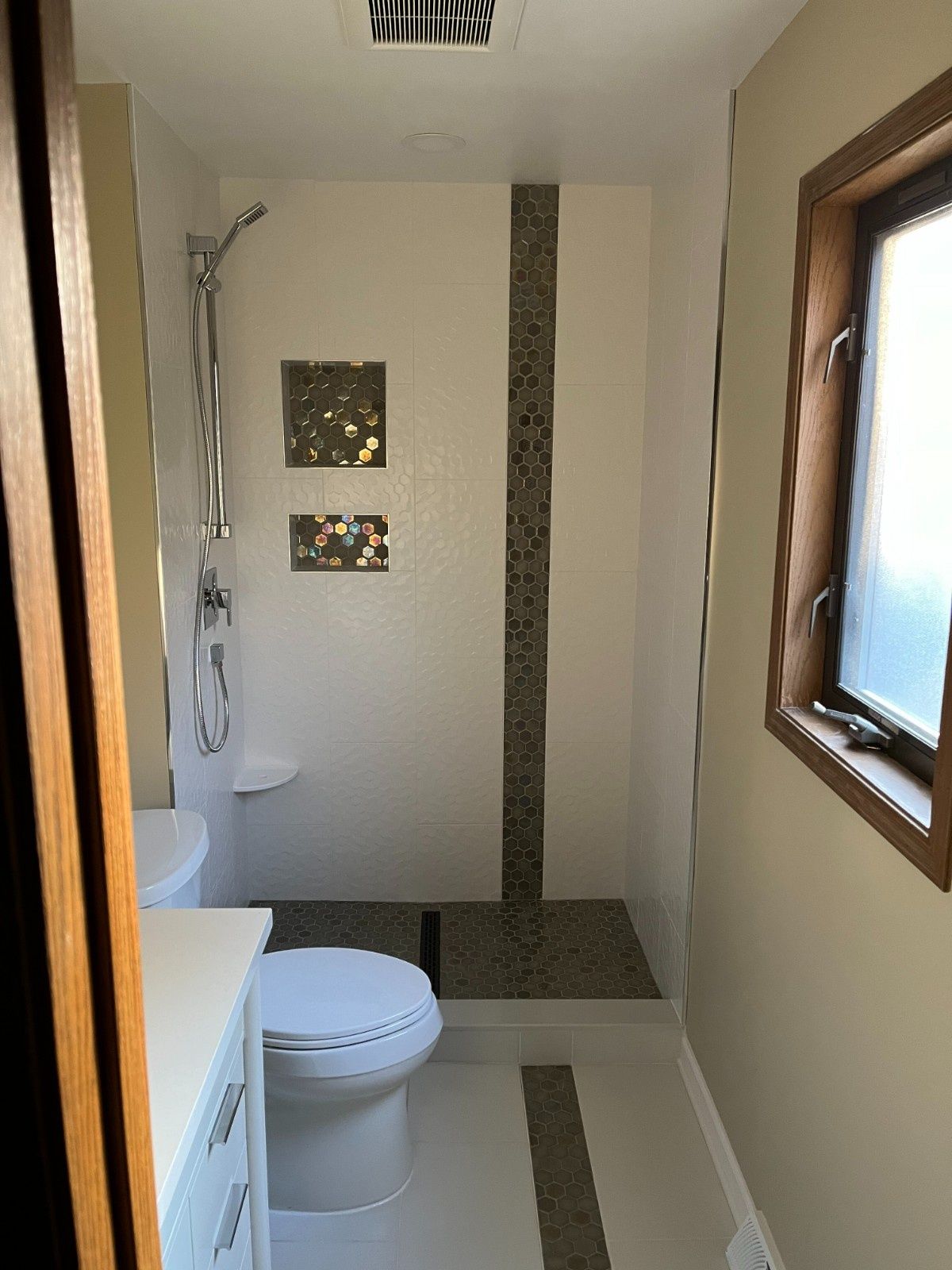 Narrow bathroom with white toilet, shower stall, and small window on the right side.