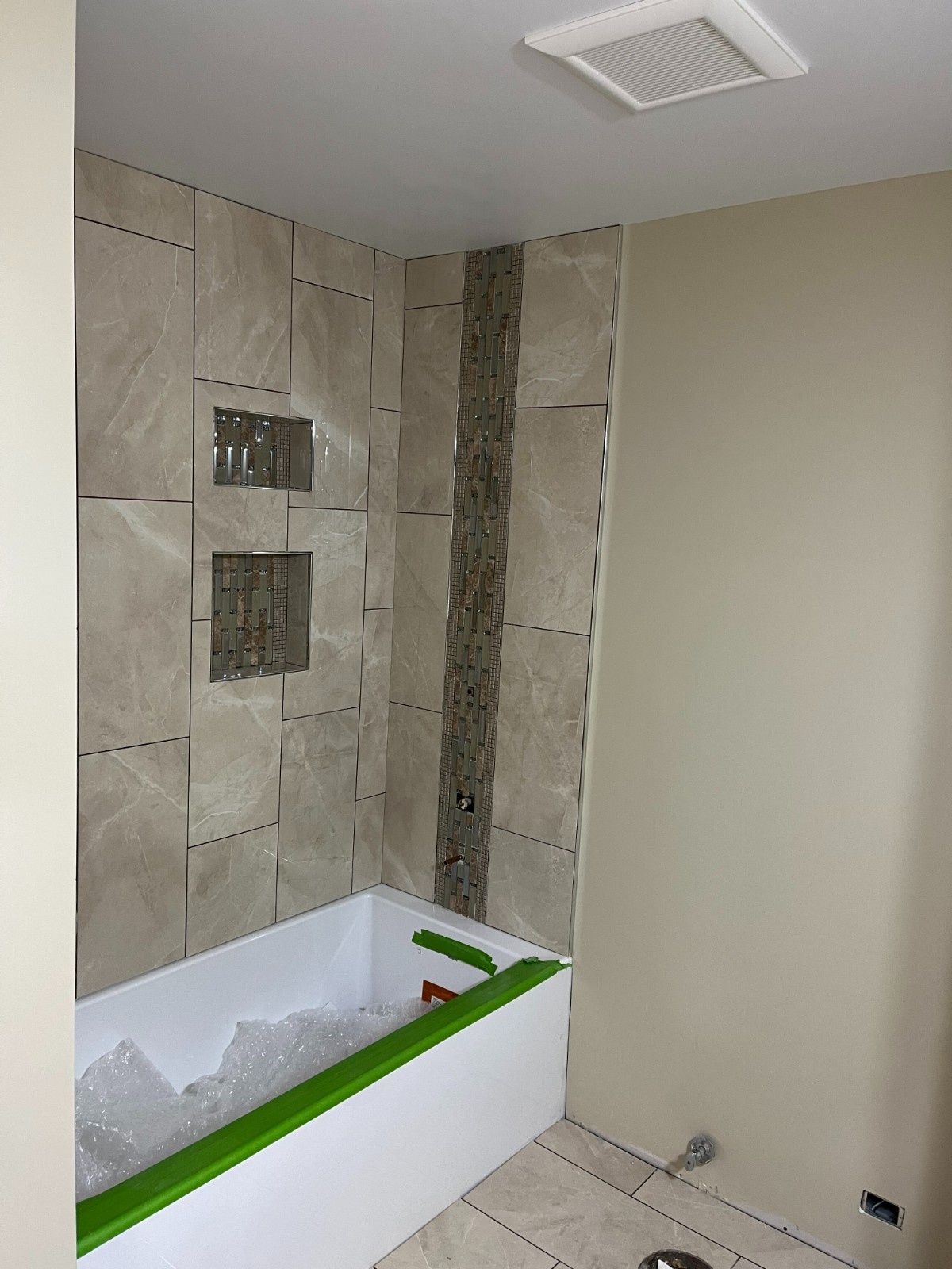 Empty bathroom under renovation with tiled shower wall and white tub surrounded by green painter’s tape