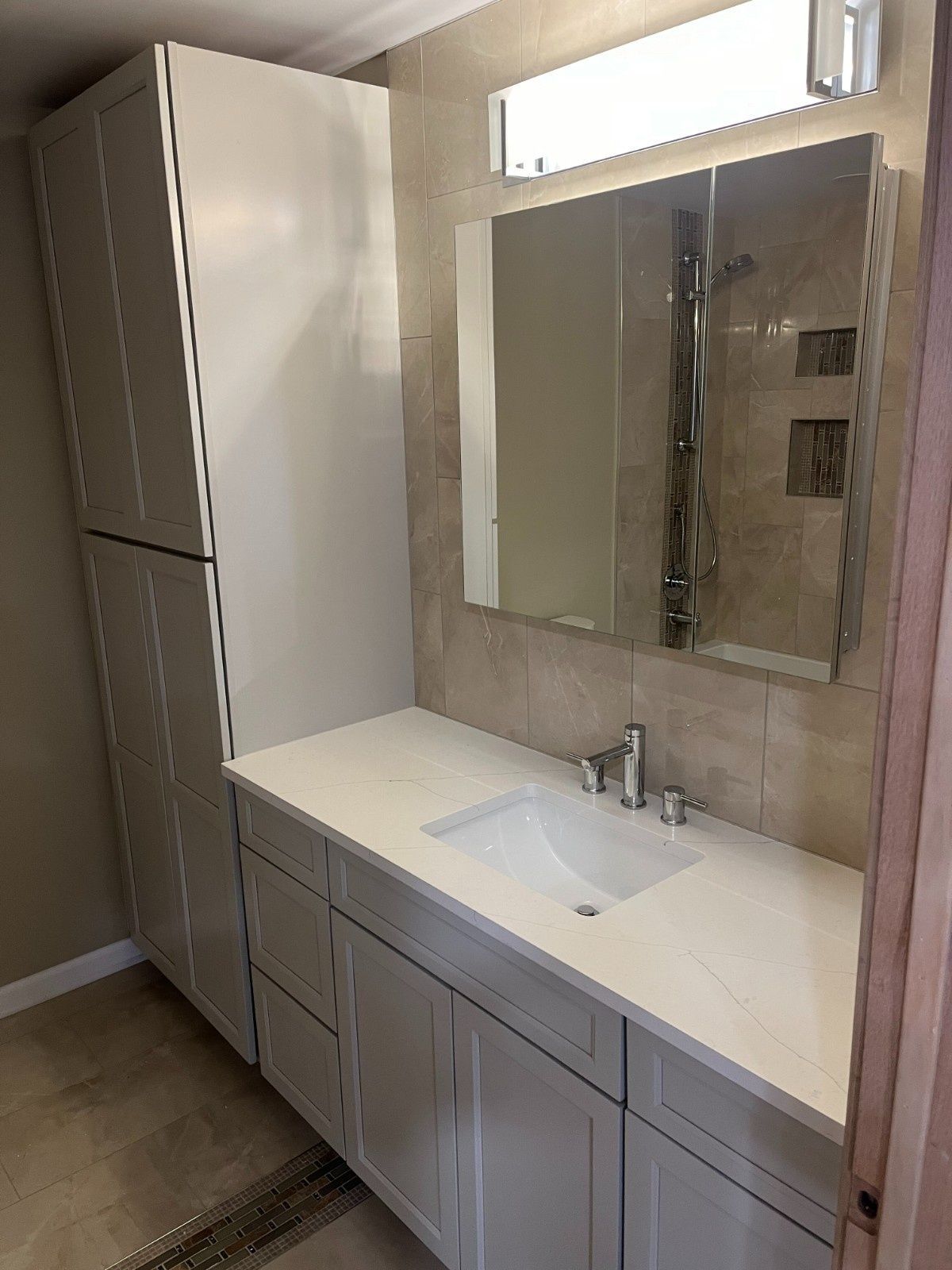 Modern bathroom vanity with white countertop, sink, mirror, and tall cabinet beside a window.