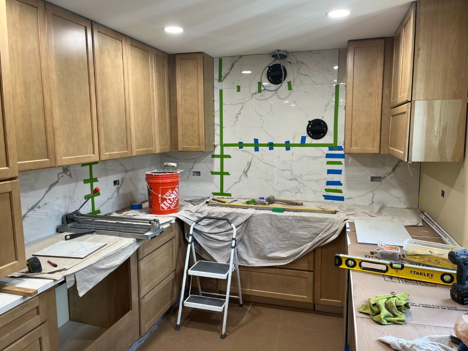 Kitchen under renovation with taped cabinets, covered counters, and tools on a step stool.