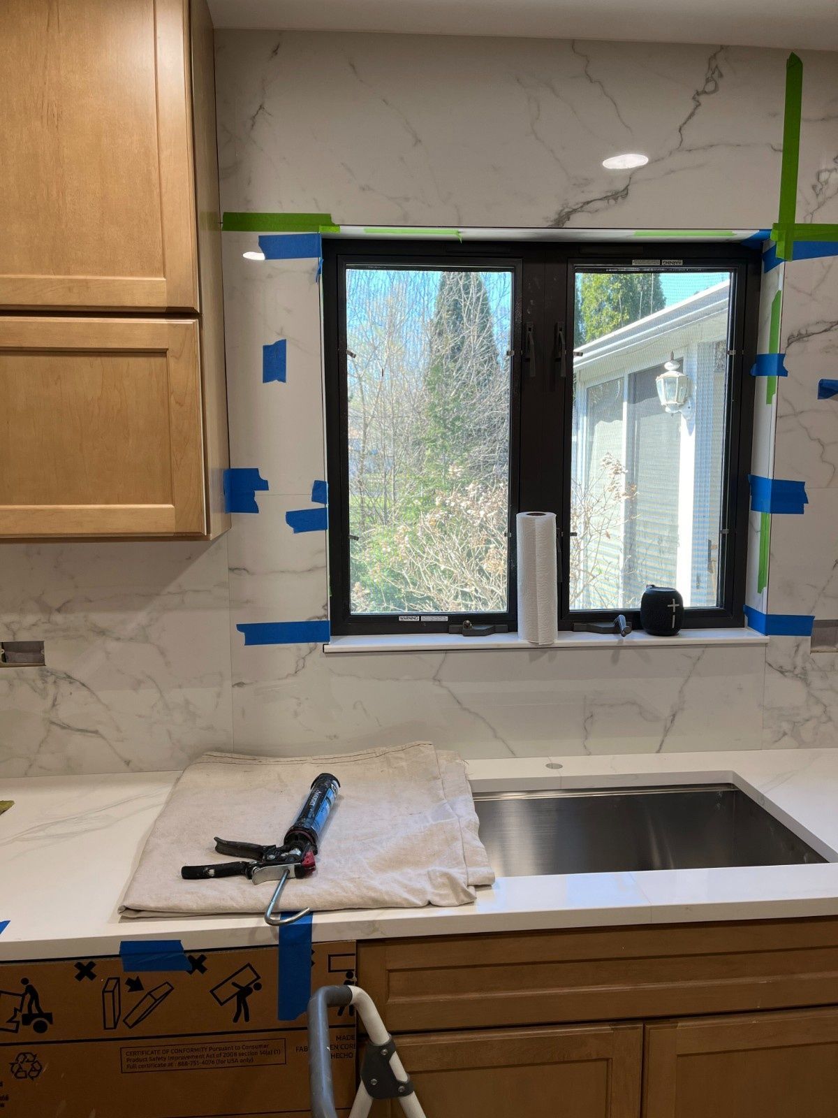 Kitchen sink and window under renovation, with blue tape, tools on the counter, and cabinets nearby