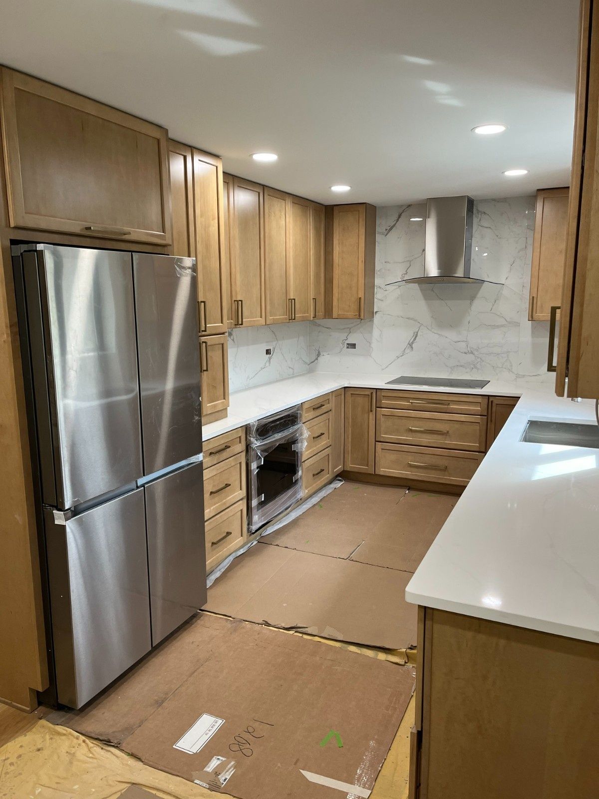 Unfinished kitchen with wooden cabinets, stainless steel fridge, white countertops, and cardboard covering the floor.