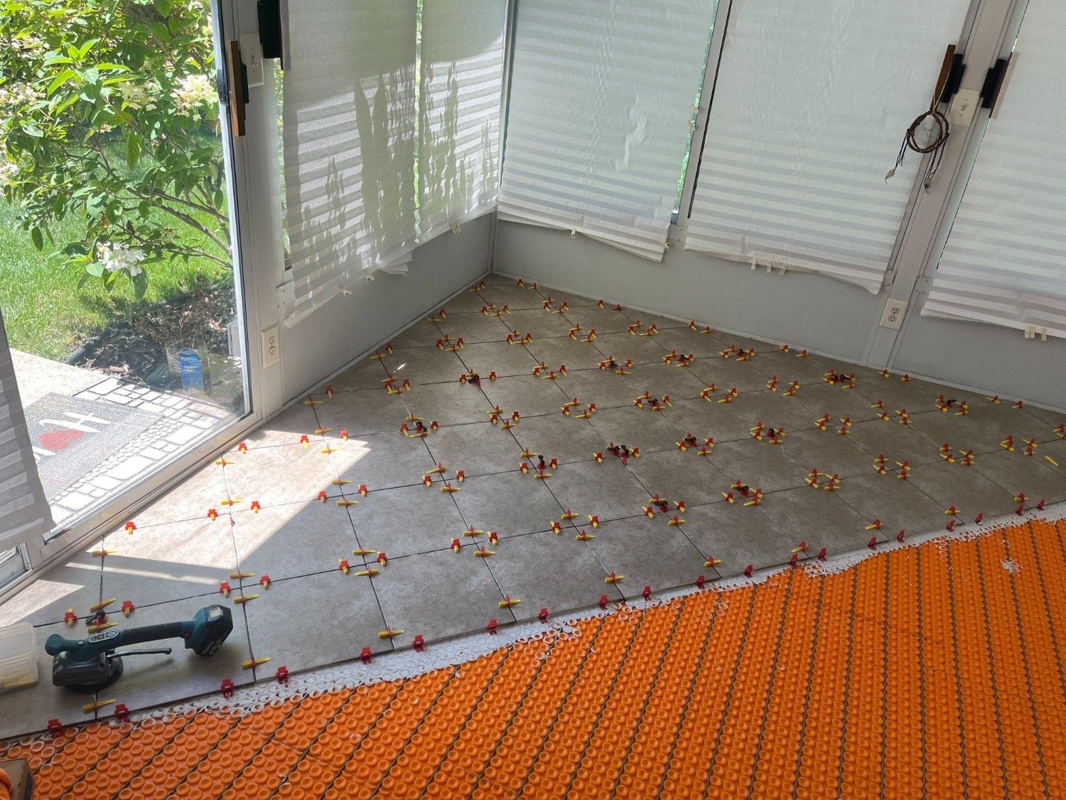 Bright sunlit room with tiled floor, window blinds, and a power tool near an orange rug edge.