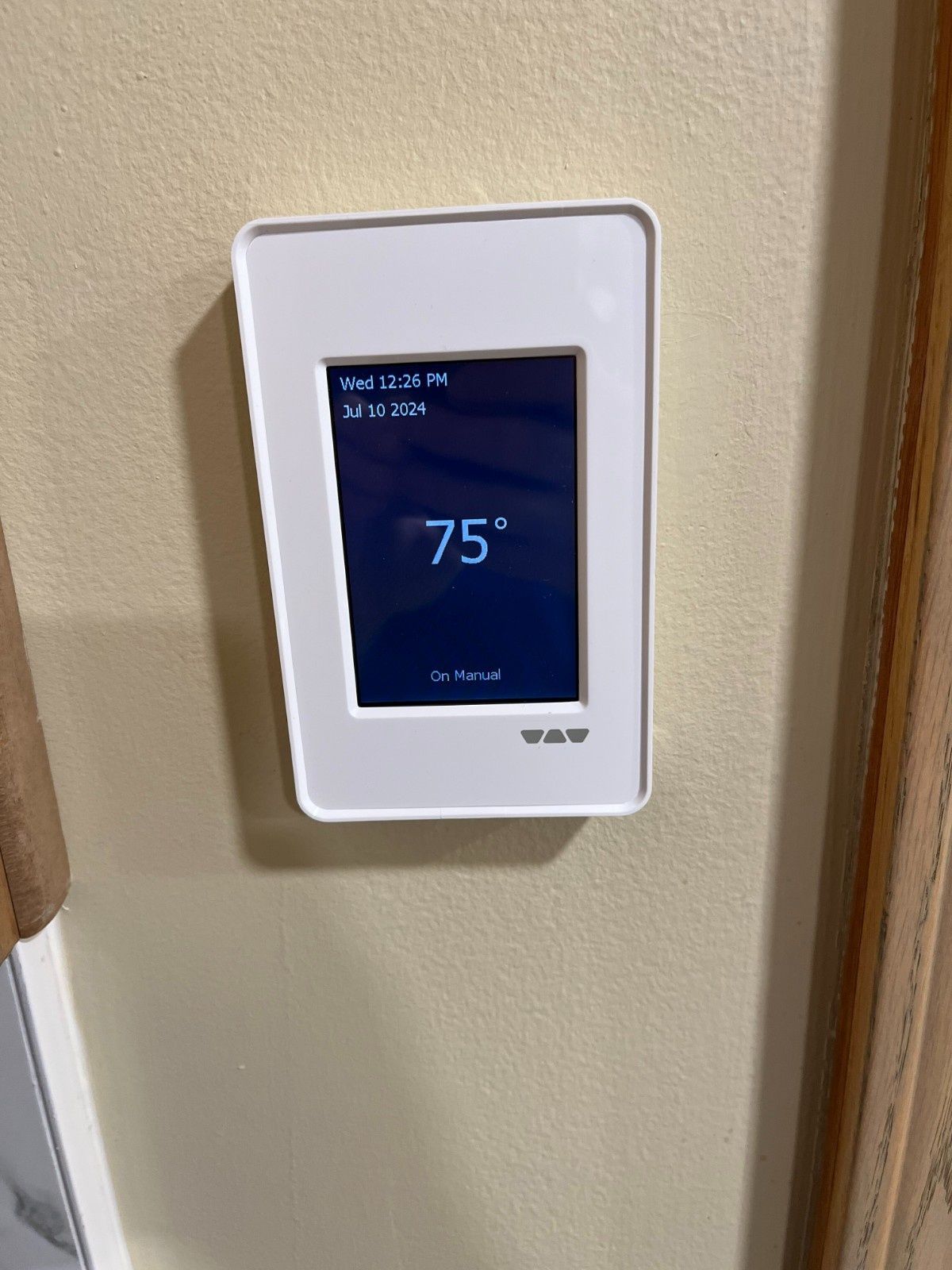 White wall-mounted thermostat displaying 75° on a beige wall near curtains