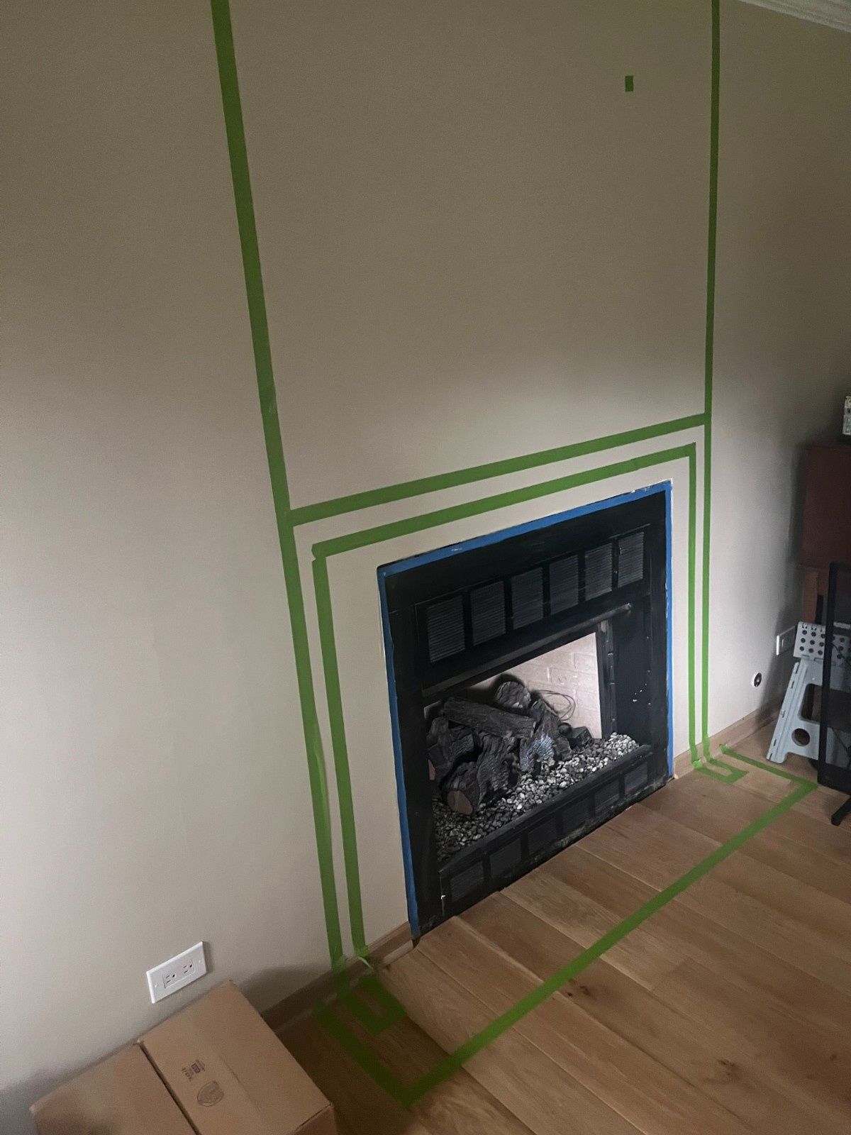 Tape-marked fireplace on a wall with hardwood floor and a cardboard box nearby.