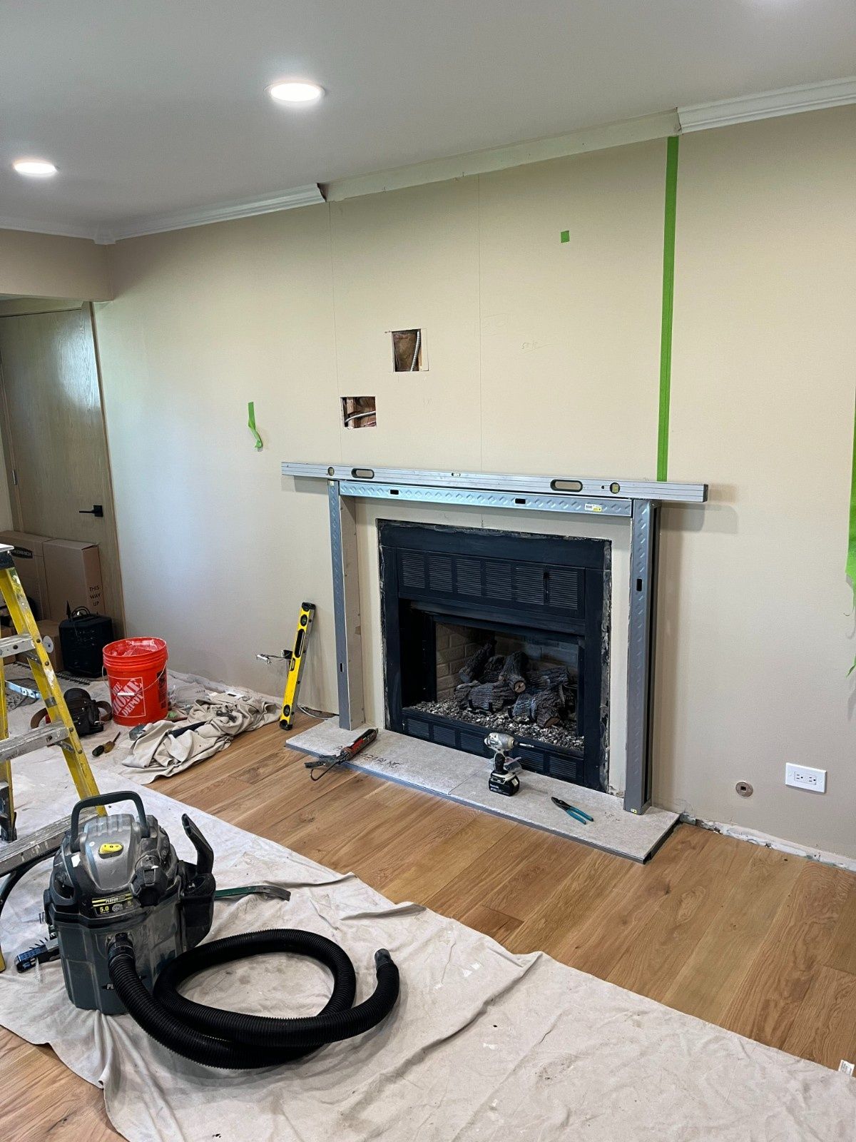 Living room with fireplace under renovation, tools and floor protection in place, wall marked for paint.