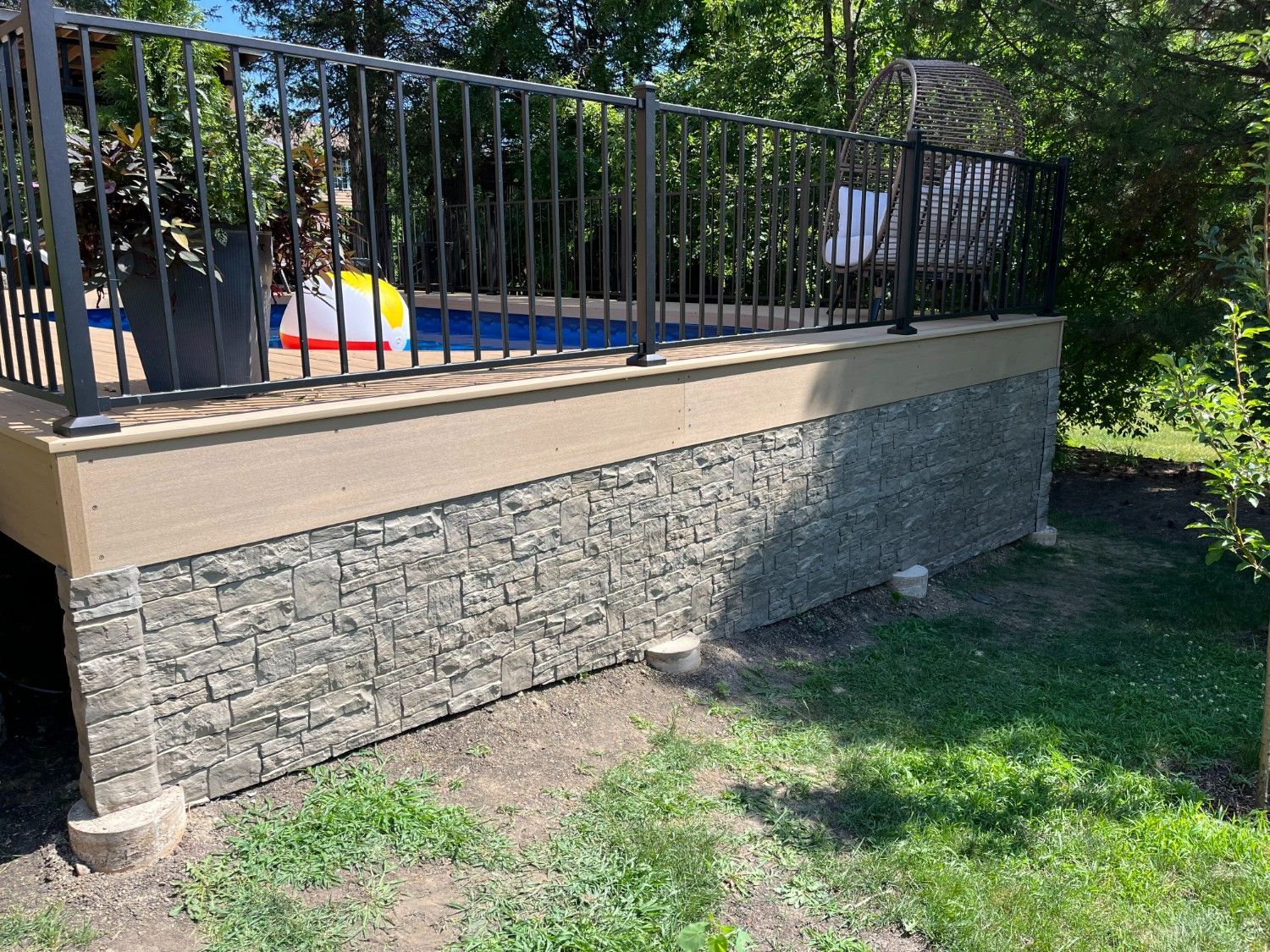 Raised stone deck with black railing beside a grassy yard and trees.